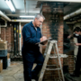 Maintenance workers in a brick basement; one on a ladder writing on a clipboard, the other wearing a mask and using a tool near a brick column.
