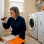 A woman uses a flashlight to inspect a wall vent in a laundry room while taking notes with an orange folder nearby and a washing machine and dryer in the background.