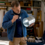 Man in a blue denim jacket inspecting a galvanized metal duct with a flashlight in a garage workshop.