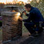 Chimney repair technician kneels on a rooftop, examining a brick chimney with a flashlight at sunset.